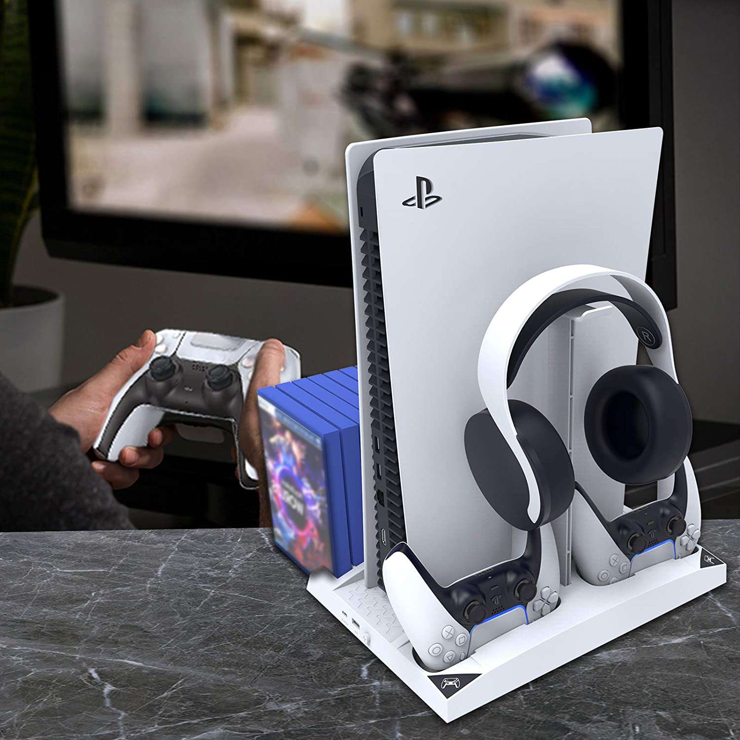 Person gaming, organized setup: PS5 controller, discs, console, headphones on charging dock